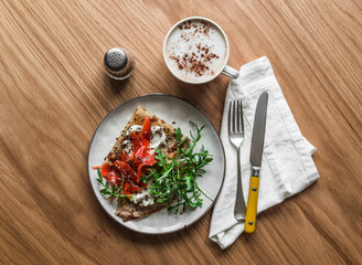 Delicious breakfast, brunch -  crepes with lightly salted salmon, cream cheese, arugula and cappuccino on a wooden background, top view
