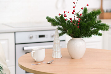Christmas kitchen interior - ceramic vase with fur branches, ceramic christmas tree, cup of tea on round wooden table in light kitchen. 