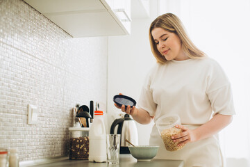 Young smiling woman in the kitchen, making breakfast from cereal and milk.