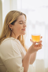 Portrait of smiling young woman holding cup of hot tea in hands in the morning in a kitchen