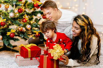 A boy together with his parents unpacks a gift box on Christmas Eve near a festively decorated Christmas tree