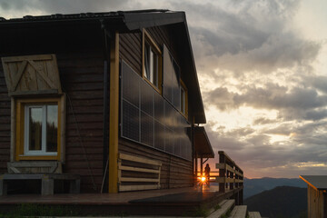 Eco-friendly cabin with solar panels on wooden wall. Unrecognizable human looking at dawn.