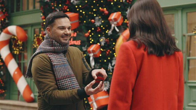 Handsome man is making a surprise proposal of marriage to his beloved woman in the winter decorated street. The woman is accepting emotionally and giving a hug full of love.