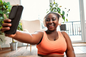 Gadgets for distance sport. Afro lady in wireless headset having video call on smartphone during daily workout at home. Happy curvy woman talking with trainer online for weight loss exercises. - obrazy, fototapety, plakaty