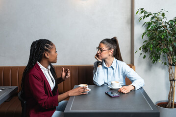Two young business woman colleagues taking a break in nearby cafeteria drinking coffee talking...