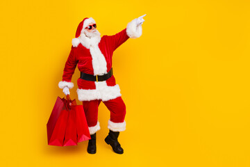 Fototapeta na wymiar Full body shot of cool santa grandfather hold red bags pointing finger empty space xmas discount offer isolated on yellow color background