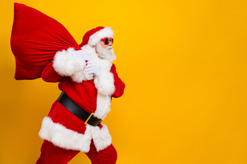 Side photo of overweight saint nicholas wear red costume walking bring huge bag with gifts for kids isolated on yellow color background