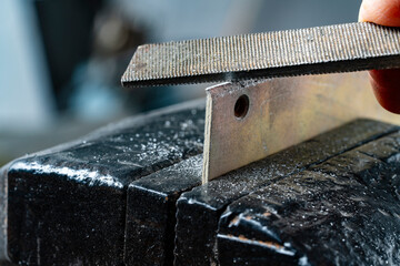 Craftsman works on small piece of metal with metal file gripped in vise on workbench table in...