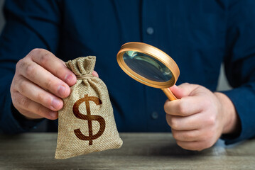 A man examines a dollar money bag through a magnifying glass. Investigating capital origins. Anti money laundering, tax evasion. Study terms and conditions on deposit or loan. Find investments - obrazy, fototapety, plakaty