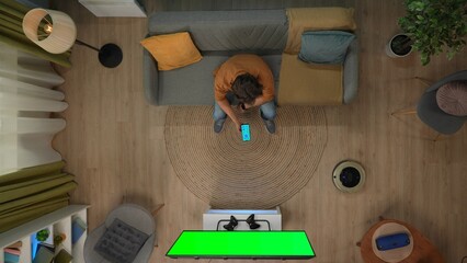 Top view of man sitting on the sofa holding smartphone looking at app for gadgets, tv with chroma...
