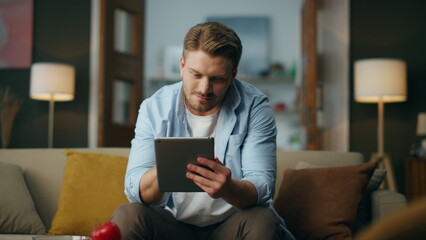 Positive man using tab computer at home. Optimistic guy working pad sitting sofa