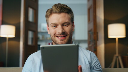 Smiling guy watching tablet screen evening room. Closeup excited man achieving