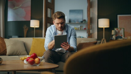 Pensive freelancer swiping tablet cozy interior. Serious guy sitting sofa alone