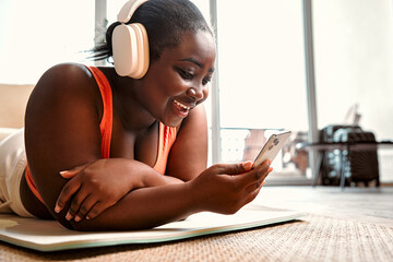 Motivation in sport. Black lady lying on stomach on yoga mat and listening music in wireless headphones. Smiling plump female in active outfit using cell phone with favorite playlist during training. - obrazy, fototapety, plakaty