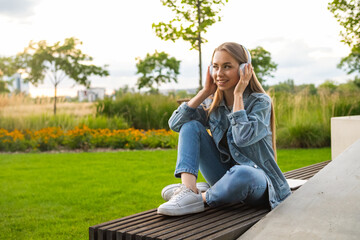 Girl with golden locks sits cross-legged on a bench, captivated by tunes playing through her...