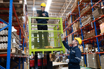 Caucasian businessman with businesswoman workers using clipboard checking Kraft paper stock while standing on scissor lift in warehouse