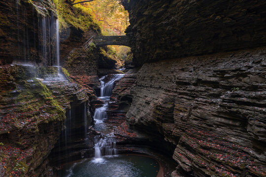 Stone bridge over river and cascades in deep limestone gorge, hiking trail going along, fallen colorful fall leafs surrounding the creek. Wide angle, long exposure. Watkins Glen State Park, NY, USA.