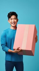 Joyful man in a blue shirt holding a large pink gift box, exuding happiness and the spirit of giving against a teal background.