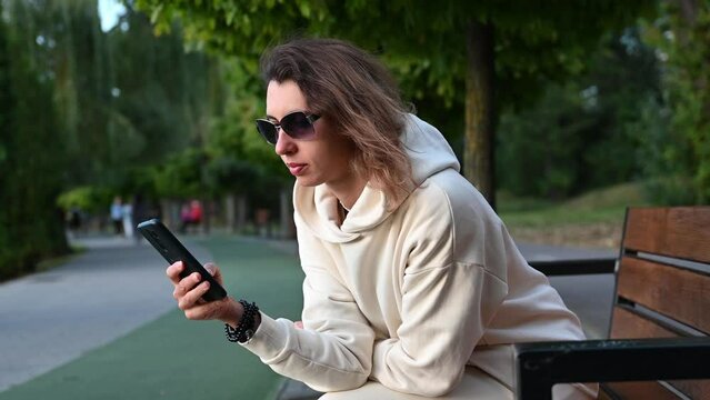 Young woman in modern clothes and sun glasses siitting on the bench in the park and scrolling social media. Relaxing in the park on sunny weather.