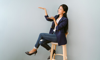 Happy cheerful Asian business woman in navy blue suit presenting or showing open hand palm with copy space for product while sitting on chair isolated on green pastel background.