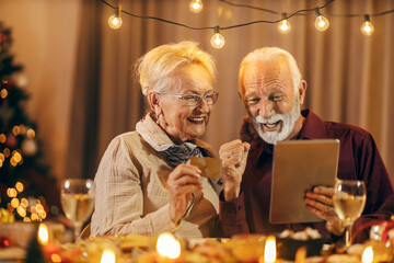 Christmas shopping. A senior couple is sitting at christmas table and using credit card and tablet for christmas shopping at home. - obrazy, fototapety, plakaty