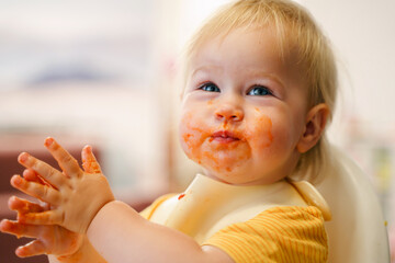 Cute toddler little child eats in a bib on a high chair, mouth dirty in sauce