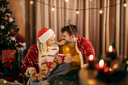 A happy couple is sitting near christmas tree at home and drinking hot beverage on christmas and new year's eve.