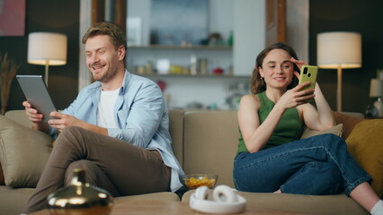 Positive couple using gadgets at home sofa. Laughing man showing tablet to wife