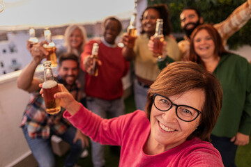 group of friends different ages party on rooftop toasting with beers, senior woman makes group selfie with smart phone