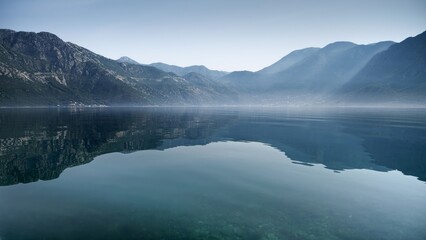 High mountains reflecting in calm sea waves with rising morning fog or mist