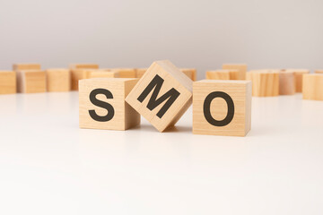 three wooden cubes with SMO symbols on them. white background. in the background there are many wooden blocks of different sizes