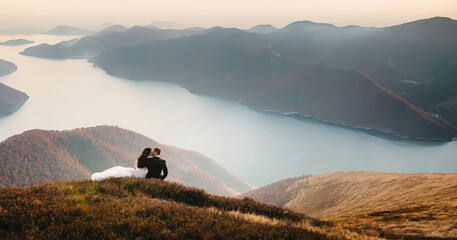 A young wedding couple is sitting on a mountain, with a mountain and a bay in the background. The...