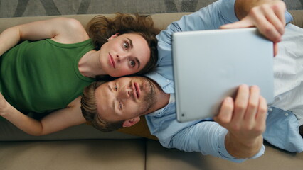 Funny couple taking selfie laying couch top view. Happy man photographing tablet