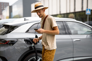 Man in hat plugs a cable in electric vehicle, while standing with phone on a public charging...