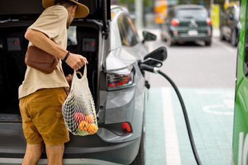 Man puts a bag full of fresh groceries in electric vehicle trunk, coming from the supermarket to...