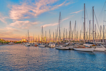 Yachts moored in the Port of Barcelona at sunset, Spain. Many boats with masts in the bay of the Mediterranean Sea against the backdrop of the city coastline illuminated by the rays of the setting sun - obrazy, fototapety, plakaty