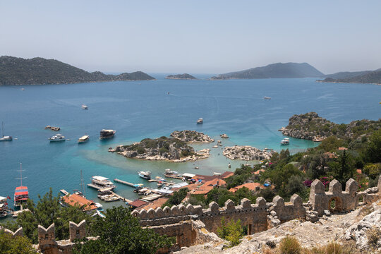 Panoramic view from the top of the Simena Castle (aka Simona Kalesi). Crenellated fortress wall along perimeter, brown tiled roofs of village houses and endless blue lagoon of the Mediterranean sea.