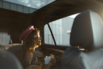 Young stylish woman sitting with a coffee cup and croissant on backseat of modern taxi while driving at city at sunny evening. Fast food and transportation concept