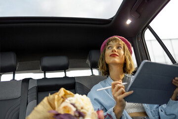 Young stylish woman uses touchpad, doing some creative work while sitting on backseat at car on the...