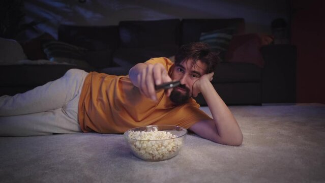 Young European man with beard lying on living room carpet eating popcorn as he changing channel smiling happily. Millennial guy enjoying movie or series and zapping with bowl inside home at night