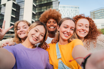 Group of young feminine women taking a selfie portrait, smiling looking at camera. Teenage girls laughing and posing for the social media photo. Multiracial excited females having fun staring front