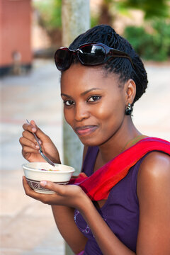 african woman with braids ,outdoors ,eating from a bowl with a spoon her breakfast