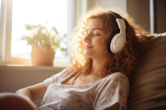 A woman enjoys the music with a headphones at the window, relaxing at home.