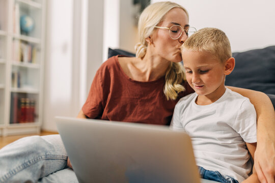 Happy mother kisses her lovely son who uses laptop.
