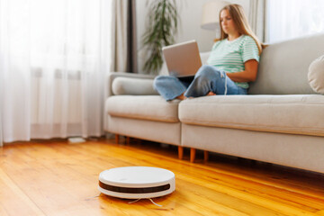 Obraz na płótnie Canvas Robotic vacuum cleaner cleaning room while woman resting on sofa using laptop
