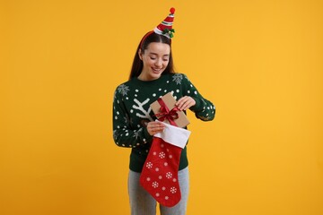 Happy young woman in Christmas sweater taking gift from stocking on orange background