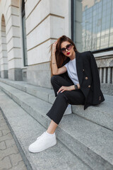 Fashionable beautiful young woman business model with vintage sunglasses in a fashion black suit with a blazer and sneakers sits on the steps near a vintage building in the city