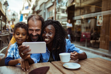 Happy multiracial couple taking selfie with little son sitting in outdoor cafe - obrazy, fototapety, plakaty