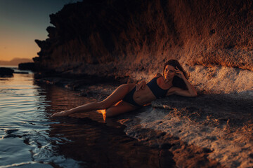 Woman in a swimsuit on the seashore with rocks at sunset