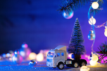 A toy truck is carrying a Christmas tree . Blue background, Christmas tree branches, balls and...